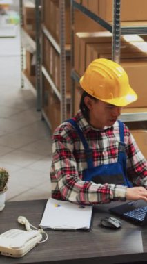 Vertical video: Asian person planning order management in warehouse, using laptop to do stock inventory. Male employee working with products in storage room, supply chain and distribution boxes.