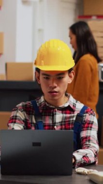 Vertical video: Asian person checking warehouse supplies from shelves, using supply chain production to work on inventory on laptop. Young man planning shipment for startup business development.
