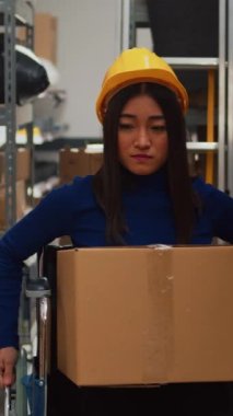 Vertical video: Asian woman in wheelchair carrying cardboard boxes to do quality control, working with employees. Female worker looking at merchandise in storage room, supply chain management.