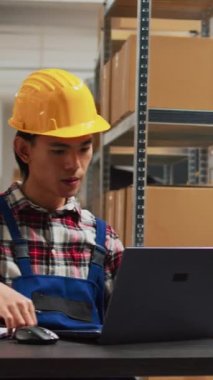 Vertical video: Young man planning order shipment in storehouse, using laptop to do stock logistics. Storage room employee working with products and merchandise, supply chain distribution packs.
