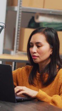 Vertical video: Female owner working in wheelchair at warehouse, using laptop to plan logistics and distribution in depot. Woman with chronic disability doing financial planning and quality control