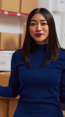 Vertical video: Young entrepreneur filming teleshopping video on phone, advertising products in warehouse space. Female worker using social media channel to create online ad promotion. Handheld shot.