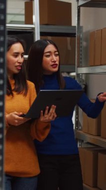 Vertical video: Asian team of people organizing products on racks, using laptop to plan stock inventory in storage room. Young women looking at boxes of merchandise on shelves, quality control.