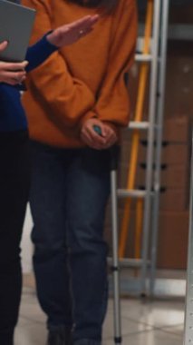 Vertical video: Female worker in crutches checking packages on racks, looking at merchandise stock in storage room and working with laptop. Team of women planning shipment for business development.