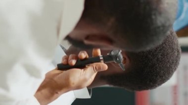 Dikey video: Eczane çalışanı otoskop aletli danışman, otolog eczane merkezinde danışmanlık ve kulak muayenesi yapıyor. Genç doktor erkek müşteriyle otoloji kontrolü yapıyor. El bilgisayarı