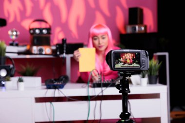 Smiling vlogger holding notebook showing product in front of camera while recording review using professional vlogging equipment. Content creator standing at desk filming podcast in broadcast studio