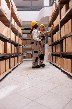 Yük dağıtım merkezi çalışanları taşımadan önce kutuları tarıyor. Koruyucu kask ve tulum giyen iki kadın depo çalışanı konuşuyor, kutularla dolu rafın yanında duruyorlar.