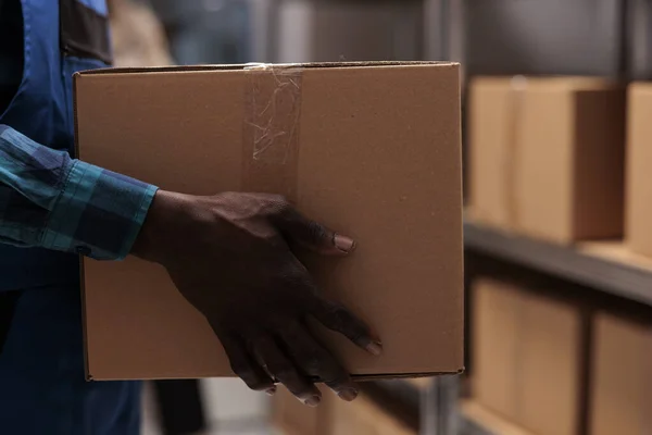 African american warehouse worker hands taking parcel from shelf. Shipping company employee working in storehouse, holding heavy cardboard box with adhesive tape close up