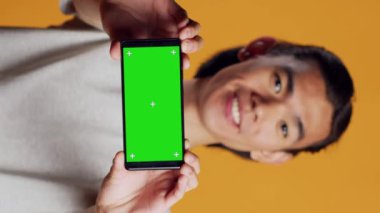 Dikey video: Erkek model yatay yeşil ekran cep telefonu tutuyor, kamerada izole edilmiş krom anahtar şablonu gösteriyor. Boş maket görüntüsü kullanarak, kopyalama ekranını kontrol ederek mutlu hisseden genç kişi