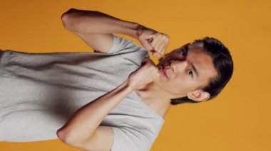 Vertical video: Furious person clenching fists and acting angry, causing conflict and being aggressive in studio. Young negative guy being violent and punching air, mad man on orange backdrop.