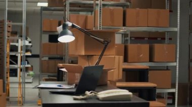 Empty storehouse with office desk to work on inventory and logistics, warehouse space filled with stacks of carton boxes on shelves. Storage room with racks, quality control on merchandise.