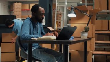 Ürünleri nakliye etmek için lojistik planlayan Afro-Amerikan işçiler, envanter bilgileriyle sipariş sevkiyatı hazırlıyorlar. Erkek sahibi dizüstü bilgisayarında ambar raflarından gelen paketlerle çalışıyor..