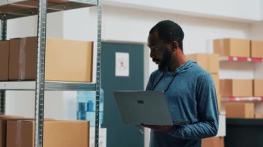 Happy person doing stock inventory with laptop in warehouse, checking goods and carton packagings before shipping merchandise. Worker looking at products, supplies management. Handheld shot.