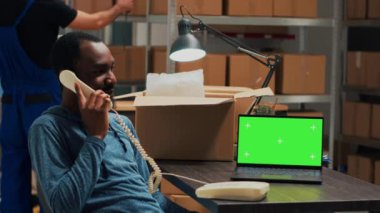 African american man talking on landline phone call and using greenscreen on laptop, working with isolated chromakey display at warehouse. Storehouse owner with telephone and mockup.