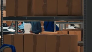 Small business owner planning supplies shipment at storehouse, putting merchandise in cardboard boxes for distribution. Man taking goods from storage shelves to ship products. Handheld shot.