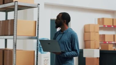 Young entrepreneur doing stock inventory with laptop, checking goods and box packagings before shipping merchandise. Male employee looking at products, supplies management. Handheld shot.