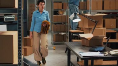 Small business owner planning order shipping with box, taking notes on laptop for stock inventory and working on delivering merchandise. Young adult selling products from warehouse racks.
