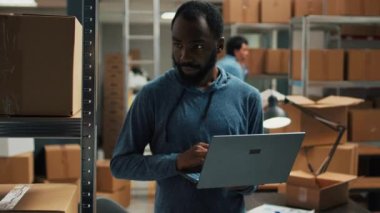 Warehouse employee using laptop to write products information, analyzing racks filled with carton packages for stock inventory. Young man planning logistics and merchandise management.
