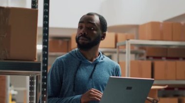 African american man working on merchandise inventory using laptop and taking notes in storehouse. Entrepreneur checking products in cardboard boxes, working on quality control and distribution.