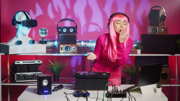 Asian performer playing electronic music at mixer console, having fun with fans in club during night time. Dj musician with pink hair mixing techno sound using professional audio equipment