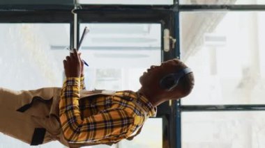 Dikey video: Depoda dans eden genç yetişkin tuhaf davranıyor, kulaklıkla neşeli şarkılar söylüyor. Modern depo çalışanı dans figürleri sergiliyor ve rafların yanında kutularla aptalca davranıyor..