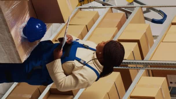 垂直ビデオ 在庫と物流に取り組んでいるアフリカ系アメリカ人従業員は 出荷のための商品を準備するためにクリップボードの在庫リストを見ています 箱の中の製品をチェックする保管室の労働者 — ストック動画