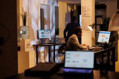 Advisor agent standing at desk table analyzing marketing statistics on computer, working at company strategy late at night in startup office. Executive manager planning investment graph