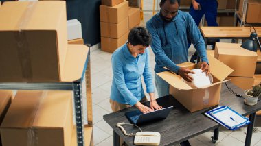 Diverse entrepreneurs giving highfive after putting products in cardboard packages, finishing quality control and delivery logistics. Man and woman feeling happy about merchandise distribution.