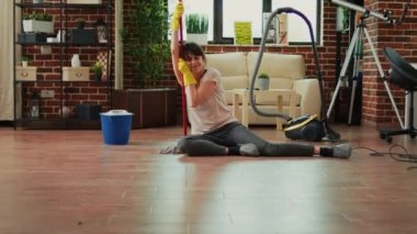 Female person feeling tired after finishing spring cleaning, sitting on clean wooden floor with mop and gloves. Young sleepy woman being proud of tidy apartment, household chores finished.