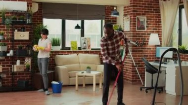 Young couple cleaning living room with vacuum and furniture polish, woman using all purpose cleaner to sweep dust from shelves. African american man vacuuming wooden floors at home.