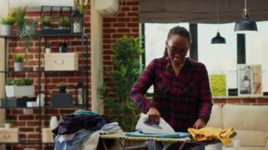 African american woman having fun ironing clothes in living room, listening to music and dancing while she does housework. Modern girl showing dance moves and being silly, enjoyment.