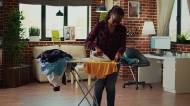 African american housewife using iron on ironing board to do housekeeping work, casual adult ironing clothes in living room. Young woman smoothing out garment and clothing, chores.