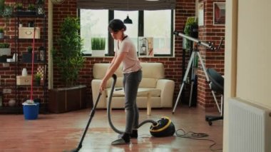 Modern housewife sweeping dust with mop in apartment, mopping floors and cleaning living room. Casual female person washing mess with cleaner, being focused on housework or chores.
