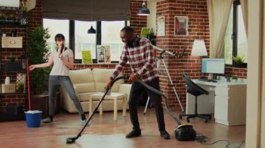 Silly couple singing song and cleaning apartment floors, using mop and vacuum cleaner at home. Diverse man and woman dancing and listening to music, washing dust and mess having fun.