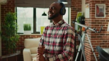 Cheerful funny boyfriend using mop to clean wooden floors, cleaning household and listening to music on headphones. Young smiling man enjoying mopping to clean dirt, household chores.