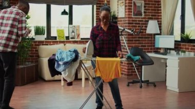 Depressed african american woman ironing laundry, getting angry at husband not helping with house chores. Tired stressed girlfriend taking care of household by herself, washed garment.