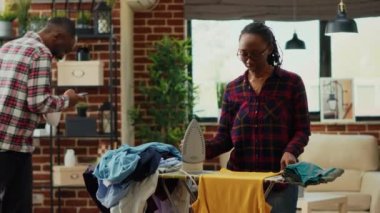African american adult using steam on ironing table to do spring cleaning, casual housewife ironing laundered clothes in living room. Young woman smoothing out garment and clothing, chores.