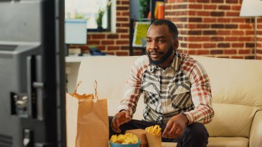 Cheerful person taking bite of cheeseburger on couch, eating burger with fries and bottle of beer. Young guy watching television and having dinner, feeling relaxed with delivery food.