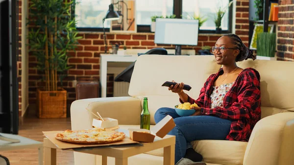 Happy woman laughing at favorite film on tv and drinking beer, holding bowl of chips in living room. Young adult enjoying movie on television with snacks and drinks, takeaway meals.
