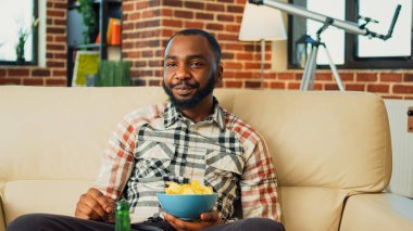 Young person drinking bottle of beer and eating snacks, enjoying favorite tv show in living room. Cheerful guy having fun with film on television, serving takeaway burgers, pizza and noodles.
