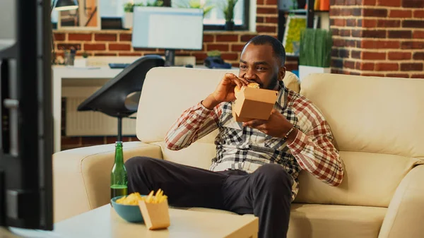 Modern guy eating cheeseburger with fries from takeout, enjoying fast food delivery meal and watching comedy movie on television. Young happy adult feeling relaxed in front of tv.