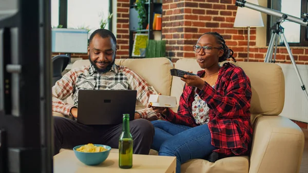 Life partners watching television together at home, eating noodles with chopsticks and browsing internet website on laptop. Cheerful modern couple having fun with tv show, drinking beer.