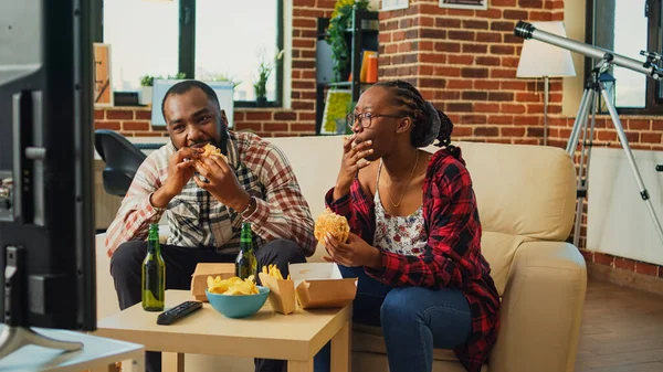 Modern life partners taking bite of cheeseburger, eating fast food from delivery takeaway at home. Young couple having dinner and enjoying comedy movie on television channel, beer bottles.