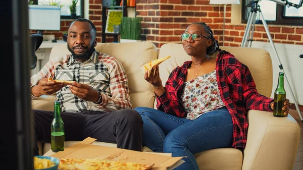 Young cheerful life partners eating slices of pizza in front of television, watching favorite movie or tv show on couch. Relaxed happy people having fast food dinner and drinking beer.