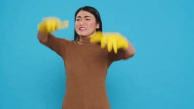 Nervous asian housekepper with protective gloves having problem with house cleaning, standing with arm crossed in studio over blue background. Maid having negative expression