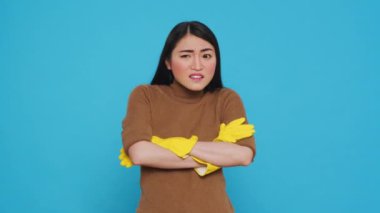 Nervous professional cleaner with protective yellow gloves having problem with house cleaning, standing with arm crossed in studio over blue background. Housekepper having negative expression