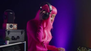 Dj with pink hair playing eletronic song at professional mixer console, listening music into headset. Asian perfomer enjoying mixing sounds, dancing with fans during night time in club.