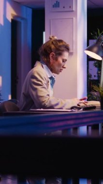 Vertical video: Corporate employee typing data on financial report, planning business development with economy graphs. Female manger checking online diagrams analytics late at night. Handheld shot.