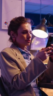 Vertical video: Young businesswoman working with smartphone at desk, using social media app while she plans financial investment late at night. Office employee analyzing diagrams. Handheld shot.