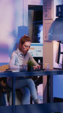 Vertical video: Young woman preparing to leave corporate job late at night, working after hours on financial growth report with marketing analytics. Business advisor leaving desk after finishing
